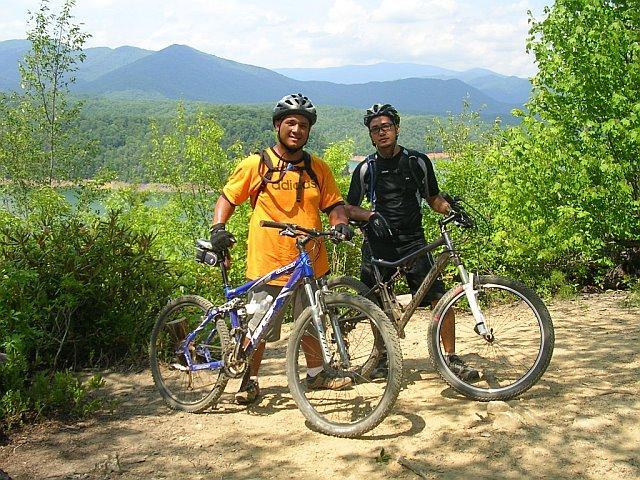 Two men pose with their mountain bikes on a dirt trail, surrounded by lush greenery and mountains in the background. The man on the left wears an orange t-shirt and a helmet, while the man on the right is dressed in a black outfit. Both are smiling and appear to be enjoying their outdoor biking adventure. Tsali Mouse Branch Loop mountain bike trail.