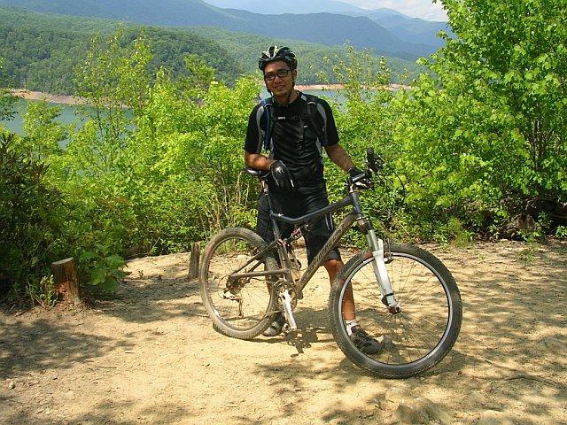 A person wearing a helmet and cycling gear stands next to a mountain bike on a sandy path, surrounded by greenery and hills in the background, with a body of water visible in the distance. Tsali Mouse Branch Loop mountain bike trail.