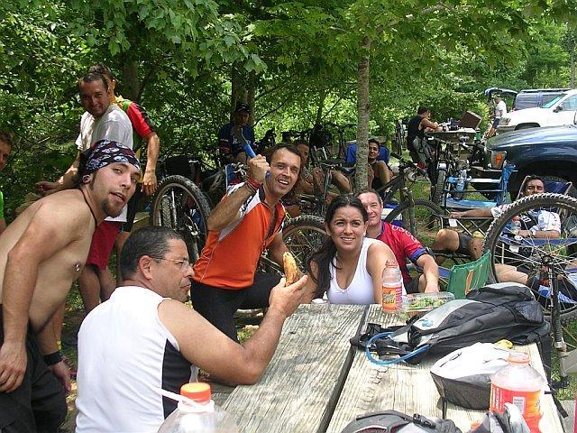 A group of mountain bikers gathered around a picnic table under trees, smiling and posing for the camera. Some are seated while others stand, with bicycles visible in the background. The atmosphere is relaxed and cheerful, showcasing a friendly camaraderie among the riders. Tsali Mouse Branch Loop mountain bike trail.
