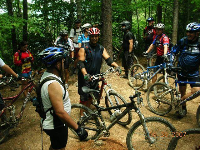 A group of mountain bikers gathered on a trail in a wooded area, wearing helmets and cycling gear. Some cyclists are adjusting their bikes while others are engaged in conversation. The atmosphere is friendly and relaxed, set against a backdrop of lush green trees. Tsali Mouse Branch Loop mountain bike trail.