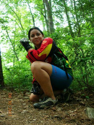 A person squatting on a forest path, wearing a colorful long-sleeve shirt and biking shorts, surrounded by green trees and foliage. The image captures a serene outdoor setting. Tsali Mouse Branch Loop mountain bike trail.