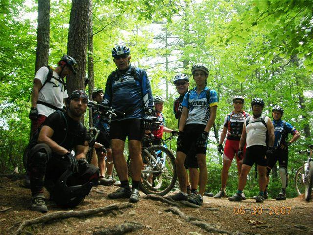 Group of mountain bikers posing near their bikes in a wooded area, surrounded by green trees. The group consists of ten riders, some wearing helmets and cycling jerseys, while others are in casual biking attire. The setting suggests a trail or outdoor adventure scene. Tsali Mouse Branch Loop mountain bike trail.