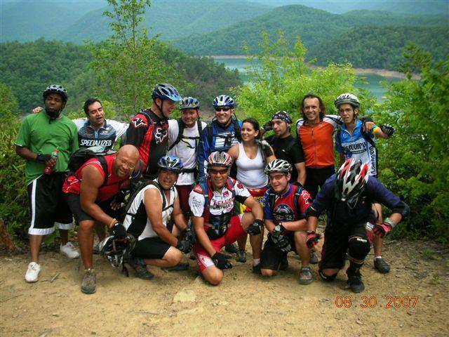 A group of 14 mountain bikers posing together outdoors, with a backdrop of green mountains and a lake. The participants are wearing bike helmets and cycling gear, and they appear to be celebrating or enjoying their time together. The setting is sunny, indicating a pleasant day for biking. Tsali Mouse Branch Loop mountain bike trail.