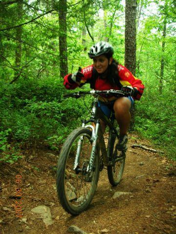 A young woman riding a mountain bike on a forest trail, wearing a helmet and a red jersey. She is focused on navigating the slightly rough terrain, surrounded by lush green trees and foliage. Tsali Mouse Branch Loop mountain bike trail.