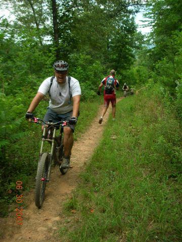A mountain biker riding on a dirt trail through a lush green forest, with a second person walking nearby. The scene captures the vibrant greenery of the surroundings and the outdoor activity of cycling and hiking. Tsali Mouse Branch Loop mountain bike trail.