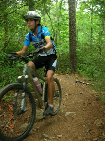 A young cyclist riding a mountain bike on a dirt trail surrounded by trees, wearing a blue and black cycling jersey. Tsali Mouse Branch Loop mountain bike trail.