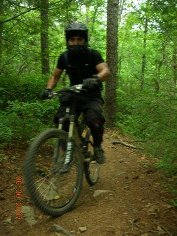 A mountain biker wearing a helmet and protective gear rides along a dirt trail surrounded by lush green trees. The biker is in motion, showcasing the rugged terrain and natural scenery of a forested area. Tsali Mouse Branch Loop mountain bike trail.