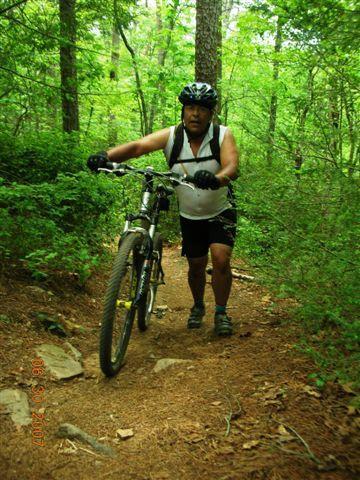A person climbing a mountain biking trail while pushing a bicycle, surrounded by lush green trees and undergrowth. The individual is wearing a helmet and athletic clothing, showcasing a determined expression. The path is made of dirt and small rocks, indicating a rugged outdoor environment. Tsali Mouse Branch Loop mountain bike trail.