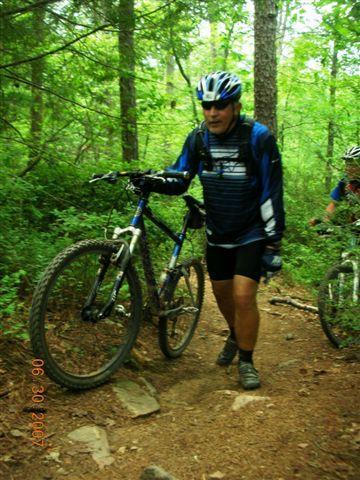 A mountain biker walking alongside their bike on a dirt trail surrounded by dense greenery. The rider is wearing a blue helmet and cycling gear, while another cyclist can be seen blurred in the background. The scene depicts a natural outdoor setting, emphasizing the adventure of mountain biking. Tsali Mouse Branch Loop mountain bike trail.
