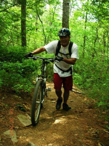 A person wearing a helmet and biking gear pushes a mountain bike up a dirt trail through a lush green forest. Trees surround the path, creating a natural, serene environment. Tsali Mouse Branch Loop mountain bike trail.