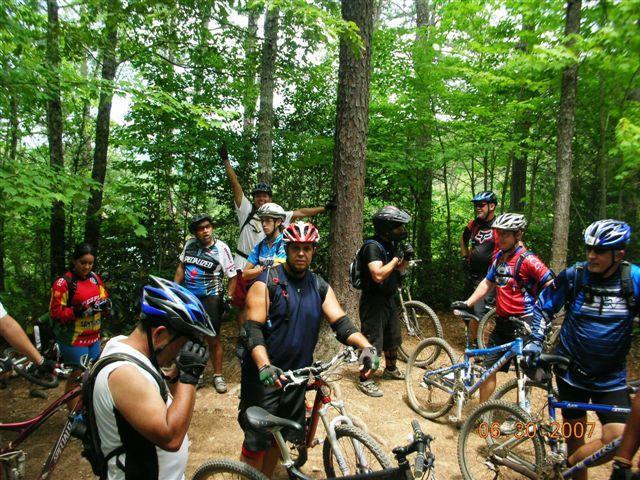 A group of mountain bikers gathered on a forested trail. Some riders are standing with their bikes, while others are adjusting their helmets or gear. The scene is surrounded by tall trees and lush greenery, indicating a lively outdoor environment. Tsali Mouse Branch Loop mountain bike trail.