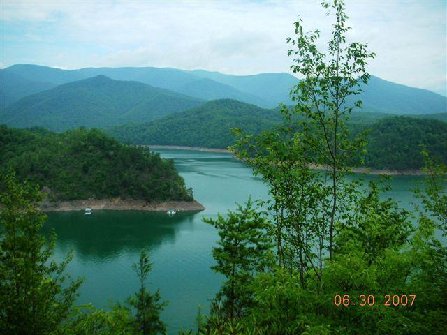 Scenic view of a tranquil lake surrounded by lush green mountains under a cloudy sky, with small boats visible on the water. Tsali Mouse Branch Loop mountain bike trail.
