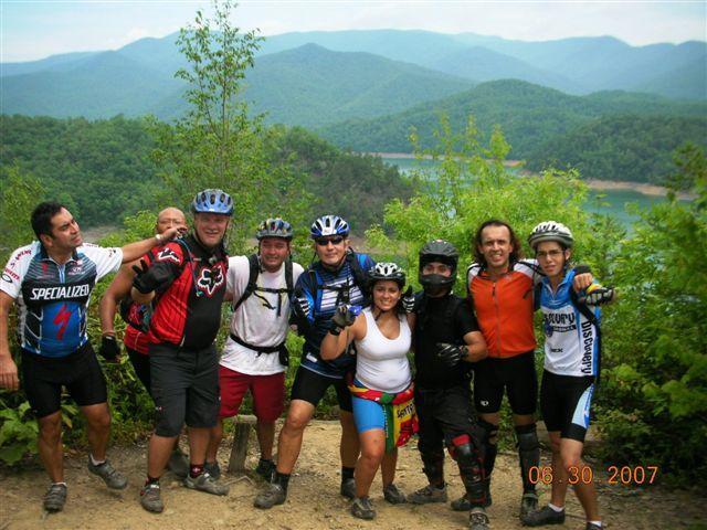 A group of eight mountain bikers poses together at a scenic overlook, surrounded by lush green mountains and a lake in the background. They are wearing various biking gear, including helmets and jerseys, and appear to be enjoying a sunny day outdoors. The image captures a sense of camaraderie and adventure among the group. Tsali Mouse Branch Loop mountain bike trail.