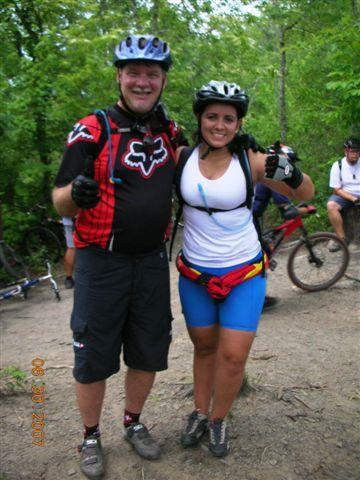 Two mountain bikers posing together in a forested area, smiling and giving thumbs up. The man is wearing a red and black cycling jersey and helmet, while the woman is in a white tank top and blue shorts with a colorful belt. Bicycles can be seen in the background, along with other riders. Tsali Mouse Branch Loop mountain bike trail.
