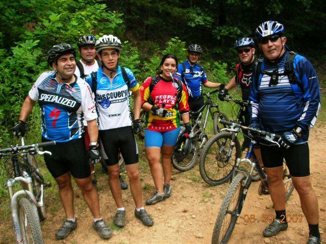 A group of seven mountain bikers posing together on a dirt trail surrounded by green foliage. They are all wearing colorful cycling jerseys and gear while standing next to their bicycles. The scene appears to be set in a natural outdoor environment. Tsali Mouse Branch Loop mountain bike trail.