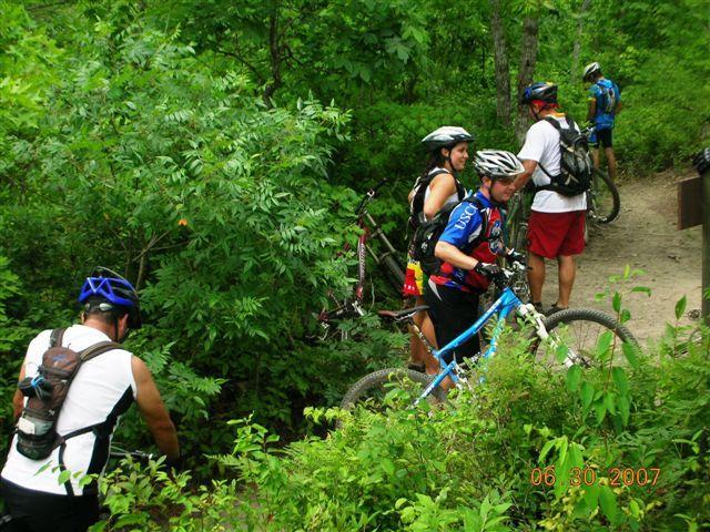 A group of five mountain bikers gathered on a lush, green trail surrounded by dense foliage. Some cyclists are adjusting their bikes, while others stand off the trail, wearing helmets and cycling gear. The scene captures the camaraderie and adventure of outdoor biking in a natural setting. Tsali Mouse Branch Loop mountain bike trail.