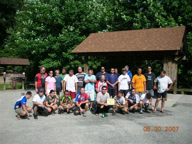 A large group of people poses for a photo outdoors in a park. They are standing and kneeling on a gravel surface, with trees and a wooden shelter in the background. The individuals are dressed casually in various sports and outdoor attire. The image is taken in daylight, suggesting a warm day, with a date marked at the bottom: June 30, 2007. Tsali Mouse Branch Loop mountain bike trail.