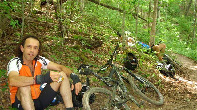 A man in an orange and white cycling jersey sits on the ground, resting while eating a snack. In the background, two mountain bikes are propped against a tree, and another cyclist is seen preparing in the woods. The scene is surrounded by lush green foliage, indicating a forested area. Tsali Mouse Branch Loop mountain bike trail.