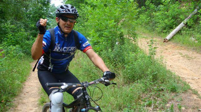 A man in a blue cycling jersey and helmet is riding a mountain bike on a dirt trail surrounded by greenery. He is smiling and raising one hand in a gesture, possibly to signal or greet someone. Tsali Mouse Branch Loop mountain bike trail.