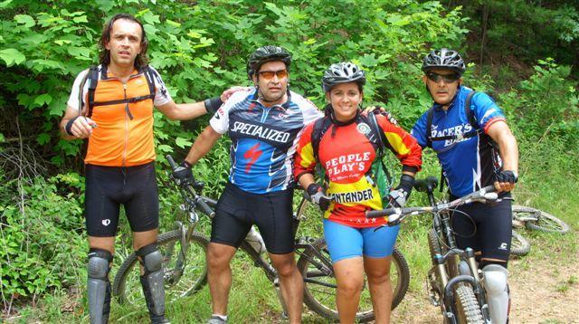 Four mountain bikers pose for a photo in a wooded area. They are wearing helmets and cycling gear, with two men on the left dressed in bright jerseys and padded shorts, and a woman in the center wearing a colorful jersey. The group appears to be taking a break during a cycling adventure, surrounded by lush greenery. Mountain bikes are leaning against them. Tsali Mouse Branch Loop mountain bike trail.