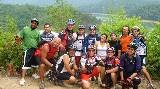 A group of diverse mountain bikers posing for a photo outdoors. They are wearing helmets and cycling gear, standing together against a scenic backdrop of trees and a lake. The atmosphere appears lively and energetic, reflecting camaraderie among the cyclists. Tsali Mouse Branch Loop mountain bike trail.