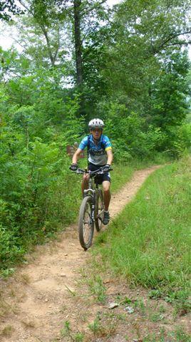 A mountain biker riding on a narrow dirt trail surrounded by lush green trees and foliage. The cyclist is wearing a helmet and cycling gear, with a focused expression as they navigate the path. The scene captures the essence of outdoor adventure and cycling in nature. Tsali Mouse Branch Loop mountain bike trail.
