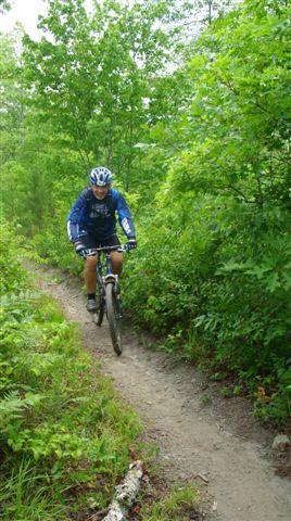 A mountain biker wearing a blue jersey and helmet rides along a narrow dirt trail surrounded by lush green foliage and trees. The trail is winding and slightly curvy, indicating an off-road biking adventure in a natural setting. Tsali Mouse Branch Loop mountain bike trail.