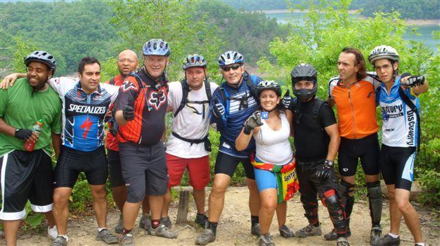 A group of ten mountain bikers posing together outdoors. They are wearing biking gear and helmets, and some are holding water bottles. The background features lush greenery and a scenic view of hills and water. The group appears to be celebrating after a ride. Tsali Mouse Branch Loop mountain bike trail.