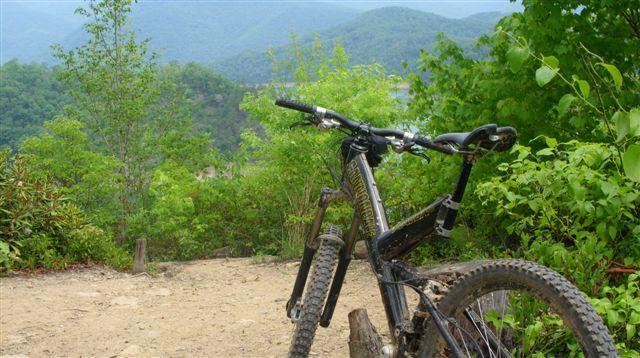 A mountain bike leaning against a wooden post on a dirt trail, surrounded by lush greenery and distant mountains under a clear sky. Tsali Mouse Branch Loop mountain bike trail.