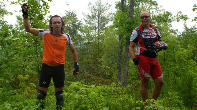 Two male mountain bikers standing in a lush green forest. One is wearing an orange jersey and black shorts, while the other is in a red and black biking outfit. They both appear to be enjoying their time outdoors, surrounded by trees and foliage. Tsali Mouse Branch Loop mountain bike trail.