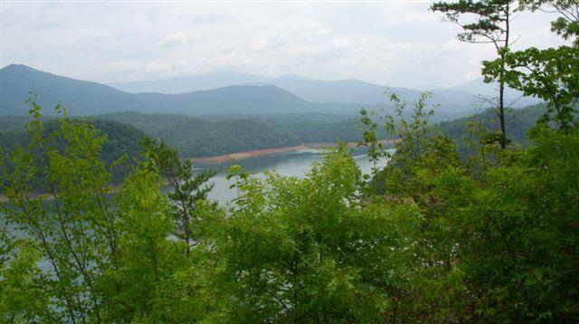 A scenic view of a mountainous landscape featuring a serene lake surrounded by lush greenery, with distant mountains under a partly cloudy sky. Tsali Mouse Branch Loop mountain bike trail.