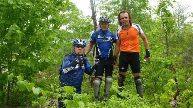 Three male mountain bikers pose outdoors among lush green foliage. They are wearing cycling gear, including helmets, with two in blue jerseys and one in orange. The group is smiling and appears to be enjoying their time. Tsali Mouse Branch Loop mountain bike trail.