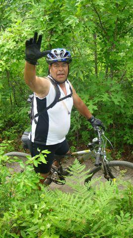 An adult male cyclist wearing a helmet and gloves is waving while standing next to his mountain bike on a trail surrounded by lush green foliage. Tsali Mouse Branch Loop mountain bike trail.