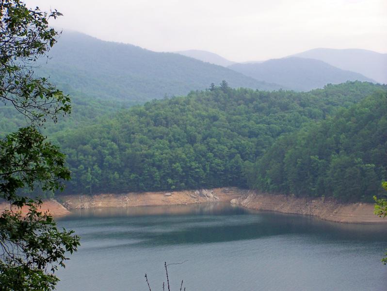 A serene landscape featuring a calm lake surrounded by lush green hills and mountains under a cloudy sky. The foreground includes foliage, while the water reflects the surrounding trees and terrain, creating a peaceful natural scene. Tsali Mouse Branch Loop mountain bike trail.