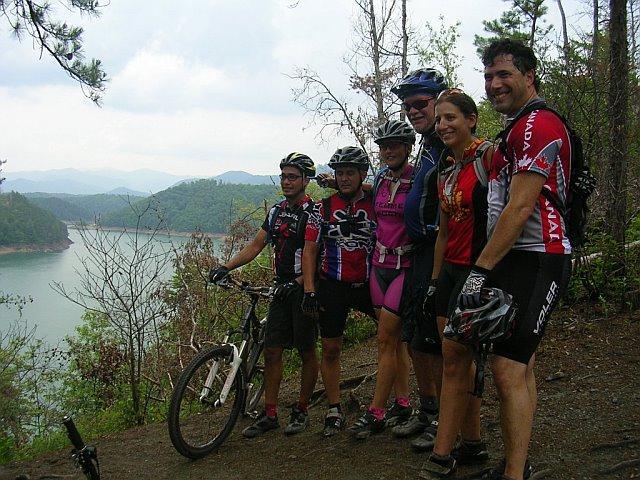 A group of six mountain bikers standing together on a scenic trail overlooking a lake, surrounded by lush greenery and rolling hills. They are dressed in cycling gear and helmets, smiling and posing for the photo. The sky is partly cloudy, enhancing the outdoor adventure atmosphere. Tsali Right Loop mountain bike trail.