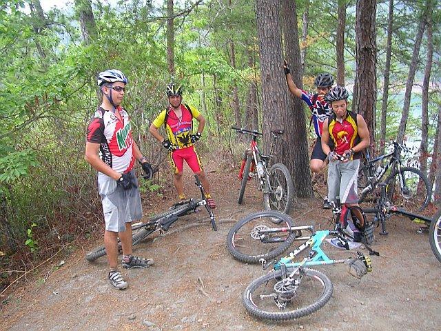 A group of four cyclists in mountain biking gear standing near their bicycles on a dirt trail surrounded by trees. The cyclists are wearing colorful jerseys, and their bikes are positioned on the ground around them. The scene captures a moment of rest during an outdoor biking activity. Tsali Right Loop mountain bike trail.