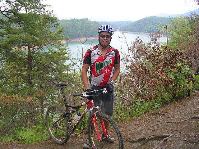A cyclist wearing a red and green branded jersey stands next to a mountain bike on a wooded trail, with a scenic view of a lake and hills in the background. The setting is lush with greenery, and the sky is overcast. Tsali Right Loop mountain bike trail.
