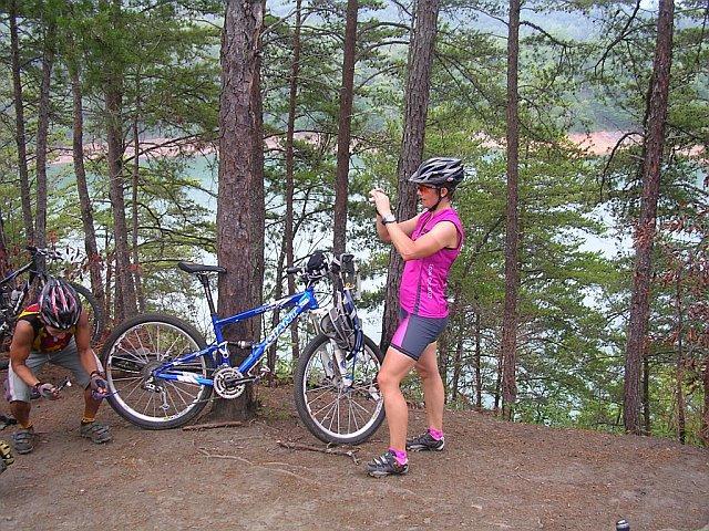 Two mountain bikers stop on a trail surrounded by trees. One biker, wearing a pink jersey and helmet, adjusts her bike while another crouches nearby, working on their gear. A scenic view of a lake is visible in the background. Tsali Right Loop mountain bike trail.