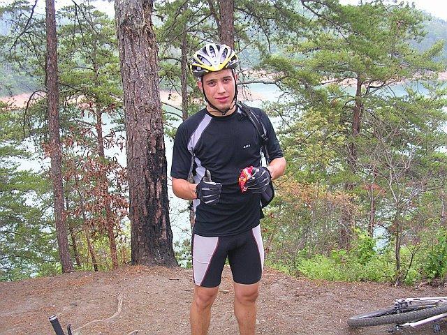 A young man in a black and gray cycling outfit and a yellow helmet stands in a forested area, holding a snack. Behind him, a scenic view of a lake surrounded by trees is visible. A bicycle is partially visible on the ground nearby. Tsali Right Loop mountain bike trail.