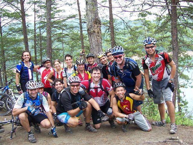 A diverse group of mountain bikers posing together in a wooded area, with trees in the background. They are wearing colorful biking jerseys and helmets, smiling and making hand gestures. The atmosphere is cheerful and energetic, showcasing a camaraderie among outdoor enthusiasts. Tsali Right Loop mountain bike trail.