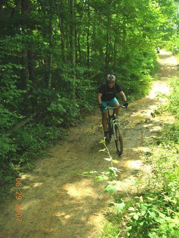 A mountain biker riding on a dirt trail surrounded by lush green trees. The cyclist is wearing a helmet and athletic gear, navigating the terrain on a sunny day. Tsali Right Loop mountain bike trail.