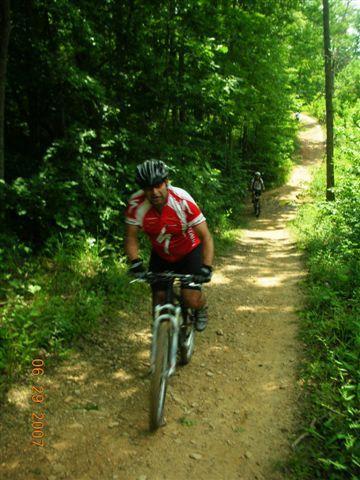 A cyclist in a red and white jersey rides a mountain bike along a dirt trail surrounded by lush green forest. Another cyclist can be seen in the background, making their way up the path. The scene captures the essence of outdoor biking adventure in a natural setting. Tsali Right Loop mountain bike trail.