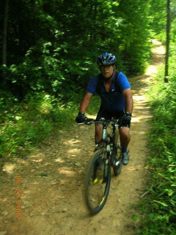 A person riding a mountain bike on a dirt trail surrounded by lush greenery. The cyclist is wearing a blue jersey and a helmet, and appears to be in motion. The setting is a wooded area with sunlight filtering through the trees. Tsali Right Loop mountain bike trail.