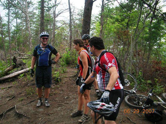 A group of four mountain bikers in a forested area, engaged in conversation and smiling. Two men and one woman are standing, wearing cycling jerseys and helmets, while a fourth person is crouched nearby with a bike. The scenery includes trees and foliage, suggesting an outdoor adventure setting. Tsali Right Loop mountain bike trail.
