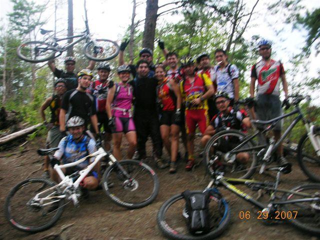 A group of mountain bikers poses for a photo in a wooded area. They are wearing colorful biking gear, and several are holding their bikes above their heads. The group is smiling and appears to be enjoying a sunny day outdoors, surrounded by trees and nature. Tsali Right Loop mountain bike trail.