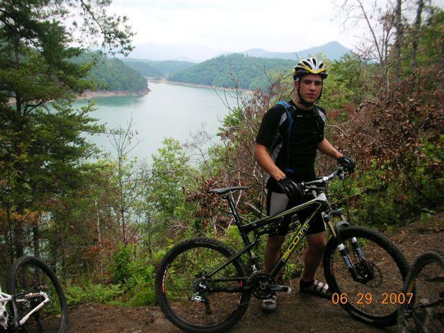 A young man in biking gear stands beside a mountain bike on a scenic trail overlooking a lake surrounded by hills. The sky is overcast, and trees are visible around the area, adding to the natural landscape. The photo is taken outdoors, capturing a moment of adventure in nature. Tsali Right Loop mountain bike trail.