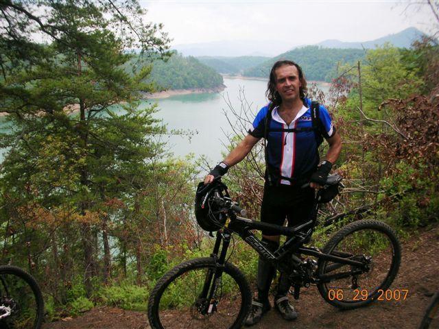 A man stands next to a mountain bike on a trail overlooking a serene lake surrounded by lush green hills. He is wearing a blue and white cycling jersey and black shorts, with his long hair flowing. The scene captures a peaceful outdoor setting, ideal for cycling and nature exploration. Tsali Right Loop mountain bike trail.