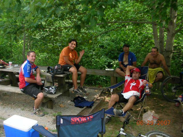 A group of five people relaxing in a shaded area after a biking excursion. They are sitting on picnic benches and folding chairs, with bicycles nearby. The scene is set in a natural environment, surrounded by greenery. One person is waving, while others are engaged in conversation. A cooler is visible in the foreground. The image captures a laid-back, outdoor gathering. Tsali Right Loop mountain bike trail.