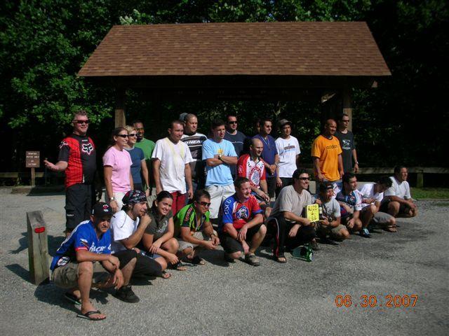 A large group of people posing together outdoors, smiling and dressed in various casual attire, including cycling jerseys and shorts. They are arranged in two rows in front of a wooden structure, with trees and greenery in the background. The image is dated June 30, 2007. Tsali Right Loop mountain bike trail.