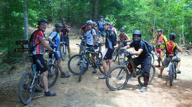 A group of mountain bikers, wearing helmets and cycling gear, gather on a dirt trail surrounded by lush green trees. They are on various bicycles, some are looking at the camera, while others appear to be chatting or preparing to continue their ride. A signpost in the background indicates the trail direction. Tsali Right Loop mountain bike trail.